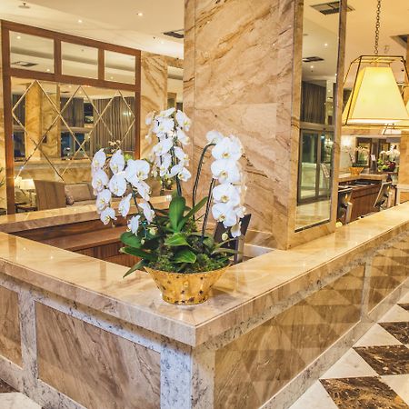 ריו דה ז'ניירו Windsor Marapendi Hotel מראה חיצוני תמונה The photo depicts a luxurious hotel lobby featuring a polished marble reception desk. There are elegant white orchids arranged in a gold pot on the counter, adding a touch of sophistication. The surrounding interior showcases marble walls, stylish li